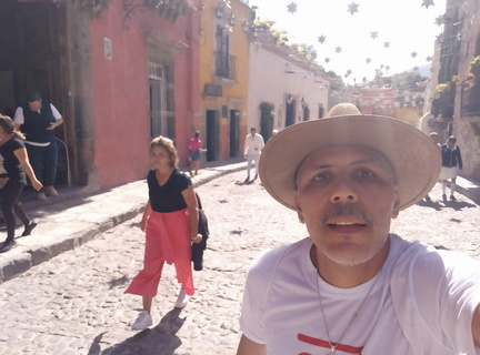 Autorretrato: Eu de chapeu e camisa branca no primeiro plano, Cida a minha esquerda de calça vermelha e blusa preta numa rua de San Miguel de Allende com suas ruass de pedras, casas coloridas e estrelas penduradas ao longo da rua enfeitando a paisagem